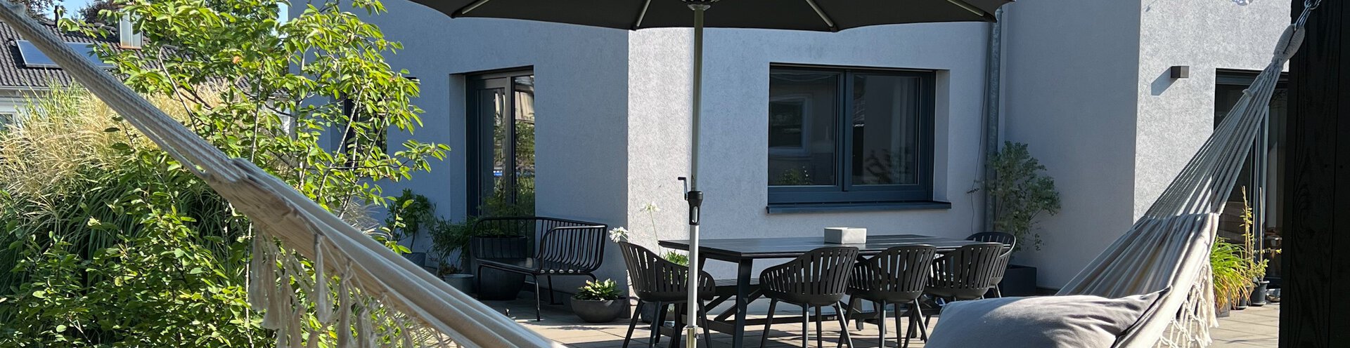 Im Vordergrund des Bildes hängt eine Hängematte, im Hintergrund steht ein Schneider Sonnenschirm des Modells Monaco auf der Terrasse vor dem Haus. Ein Tisch mit Stühlen ergänzt die gemütliche Gartenszene – ideal für entspannte Stunden im Schatten.
