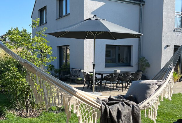 Im Vordergrund des Bildes hängt eine Hängematte, im Hintergrund steht ein Schneider Sonnenschirm des Modells Monaco auf der Terrasse vor dem Haus. Ein Tisch mit Stühlen ergänzt die gemütliche Gartenszene – ideal für entspannte Stunden im Schatten.