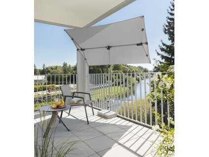 Schneider Sonnenschirm Novara in Silbergrau, 190x140 cm, auf einem Balkon mit Metallgeländer. Im Schatten des Schirms stehen ein Stuhl und ein kleiner, runder Tisch mit einem Obstteller. Auf dem Stuhl liegt ein aufgeschlagenes Buch. Im Hintergrund ein Fluss, gesäumt von vielen Bäumen.