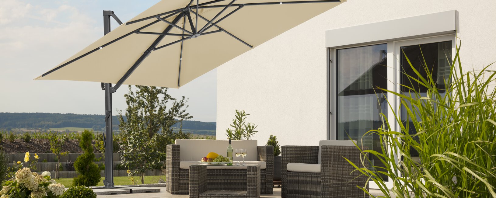 Schneider Sonnenschirm Rhodos Smart in Natur, 300x300 cm, auf einer Terasse, darunter Lounge-Möbel. Auf dem Tisch eine Obstschale, eine Flasche und Gläser. Im Hintergrund eine gestufte Stützmauer aus Betonsteinen. Um die Terrasse Wiese und Sträucher.